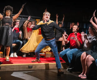 Improv actor with a yellow cape and mustache strikes a dramatic pose on a small red-carpeted theater stage as an enthusiastic audience around the stage raises hands and cheers.