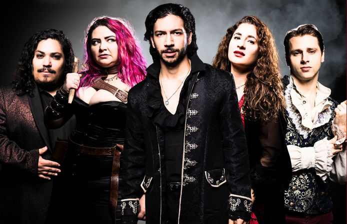 Five-person fantasy band in a dramatic studio portrait: lead in an ornate black coat, pink-haired corseted woman holding a wooden stake, two members in embroidered vests and one in a patterned jacket against a smoky backdrop.