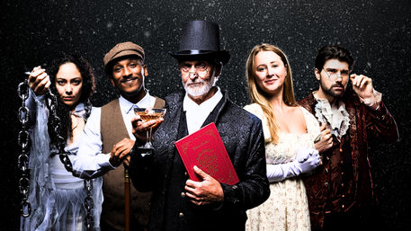 Victorian-era theater ensemble in falling snow: five costumed actors — a ghostly woman with heavy chains, a smiling man in a cap holding a cane and champagne coupe, a bearded gentleman in a top hat clutching a red book and goblet, a woman in a Regency-style white dress and gloves, and a dashing man in an ornate coat with a monocle — holiday period-costume portrait.