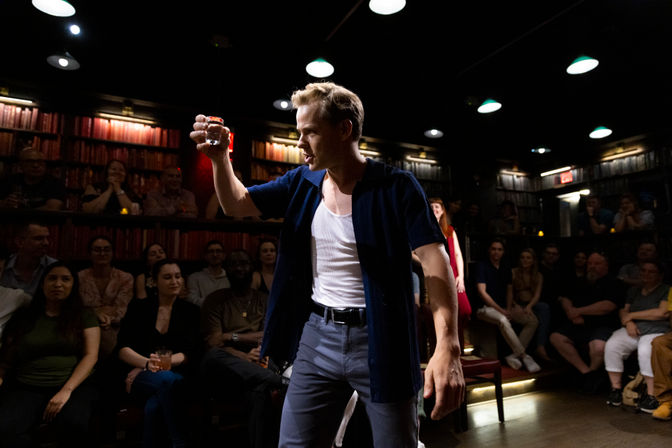 Actor in an intimate speakeasy-style theater raises a shot glass toward an attentive, closely seated audience under moody overhead lights