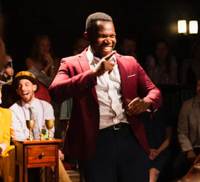 Smiling performer in a maroon blazer gestures on stage during a lively improv comedy show in an intimate club with a laughing audience
