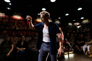 Actor raising a shot glass mid-performance in an intimate theater-bar, surrounded by a seated audience and warm bookshelf-lit backdrop