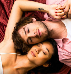 Romantic couple close-up on red patterned bedding, heads touching; man with stubble and wrist tattoo, woman with eyes closed and red-painted nails.