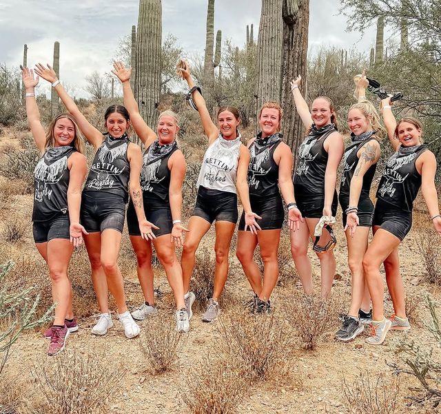 Eight women in matching black tops, bandanas, athletic shorts and sneakers pose with arms raised, smiling during a celebratory desert hike among tall saguaro cacti in the Sonoran Desert, Arizona.
