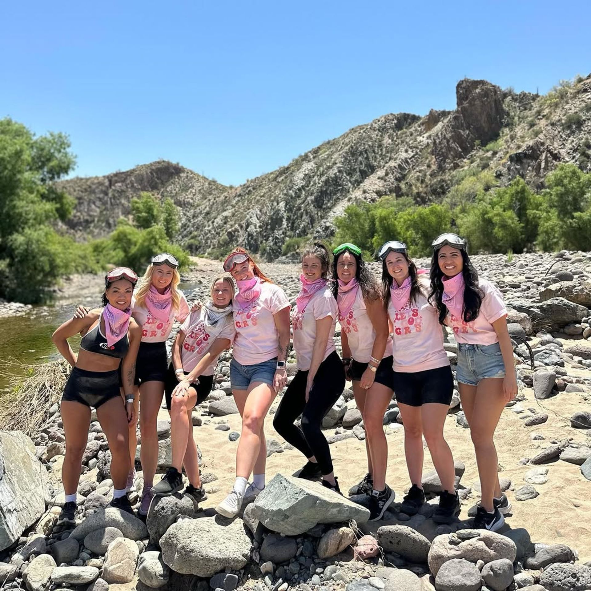 Group of eight women wearing pink bandanas and goggles posing on a rocky riverbed in a sunlit desert canyon with green trees and rugged hills