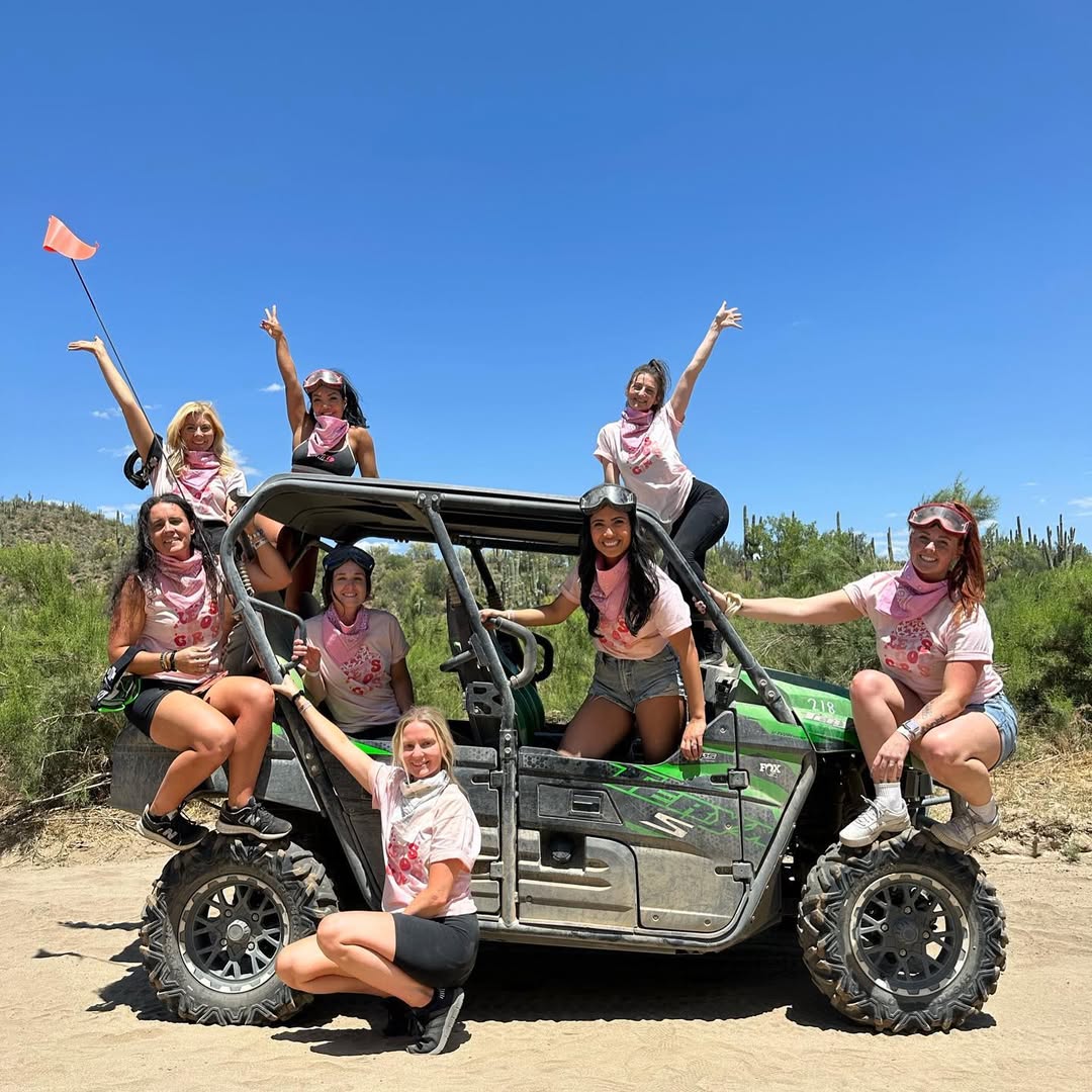 Eight women in pink bandanas posing on a green off-road UTV on a sunny Arizona desert trail with saguaro cacti and clear blue sky.