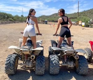 Two people in sporty outfits and sunglasses seated on off-road ATVs on a sunlit desert trail with blue sky and low scrubby hills in the background.