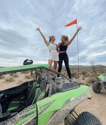 Two women standing on top of a muddy green off-road dune buggy celebrating with arms raised on a desert trail, scrub brush and distant mountains under a cloudy sky with an orange safety flag.