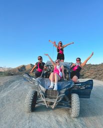 Four friends wearing matching pink accessories and sunglasses posing on a dusty off‑road UTV in a sunlit desert landscape, arms raised and smiling under a clear blue sky.