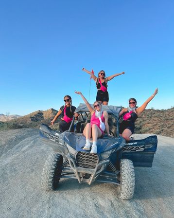 Four friends wearing matching pink accessories and sunglasses posing on a dusty off‑road UTV in a sunlit desert landscape, arms raised and smiling under a clear blue sky.