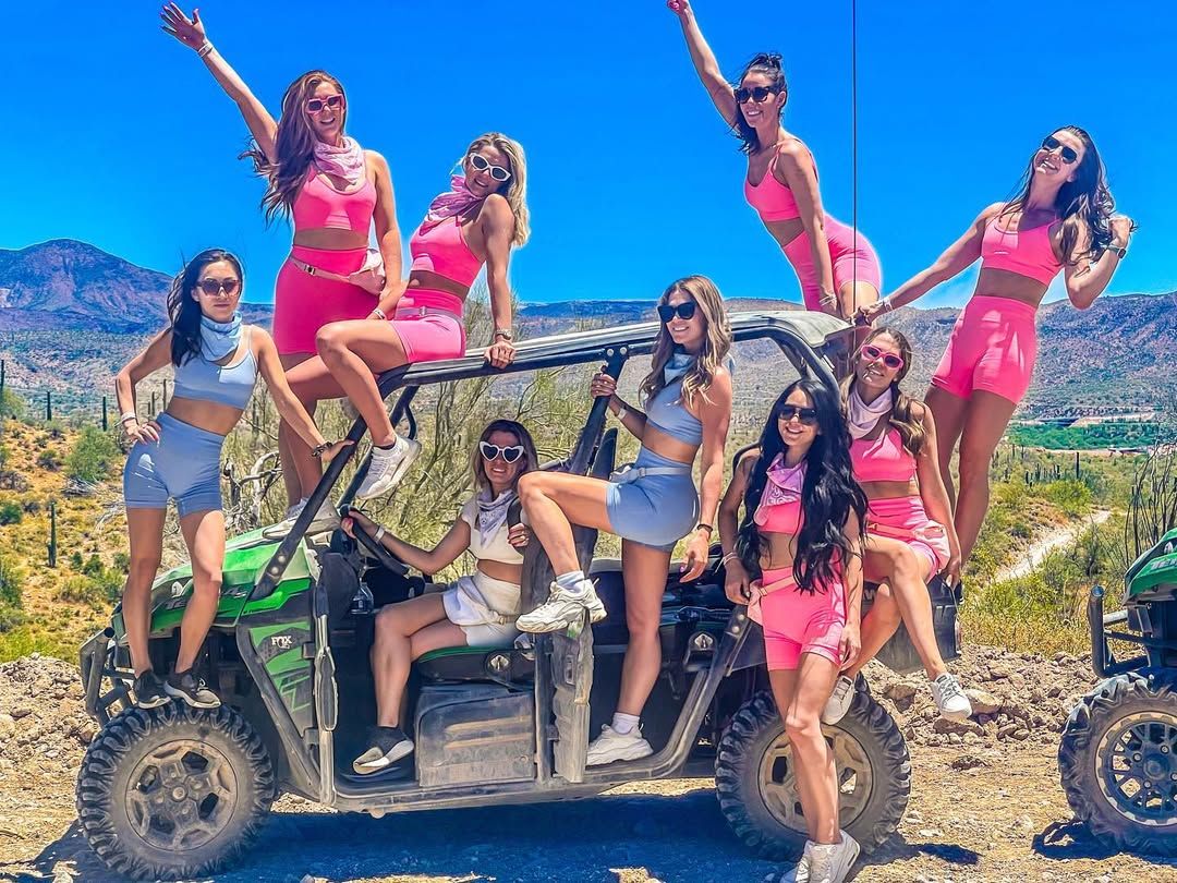 Cheerful group of women in bright pink and blue activewear and sunglasses posing on a green off-road UTV in a sunny desert mountain landscape with cacti