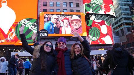 Three people in winter coats smiling and waving in a crowded NYC billboard plaza beneath colorful holiday LED screens