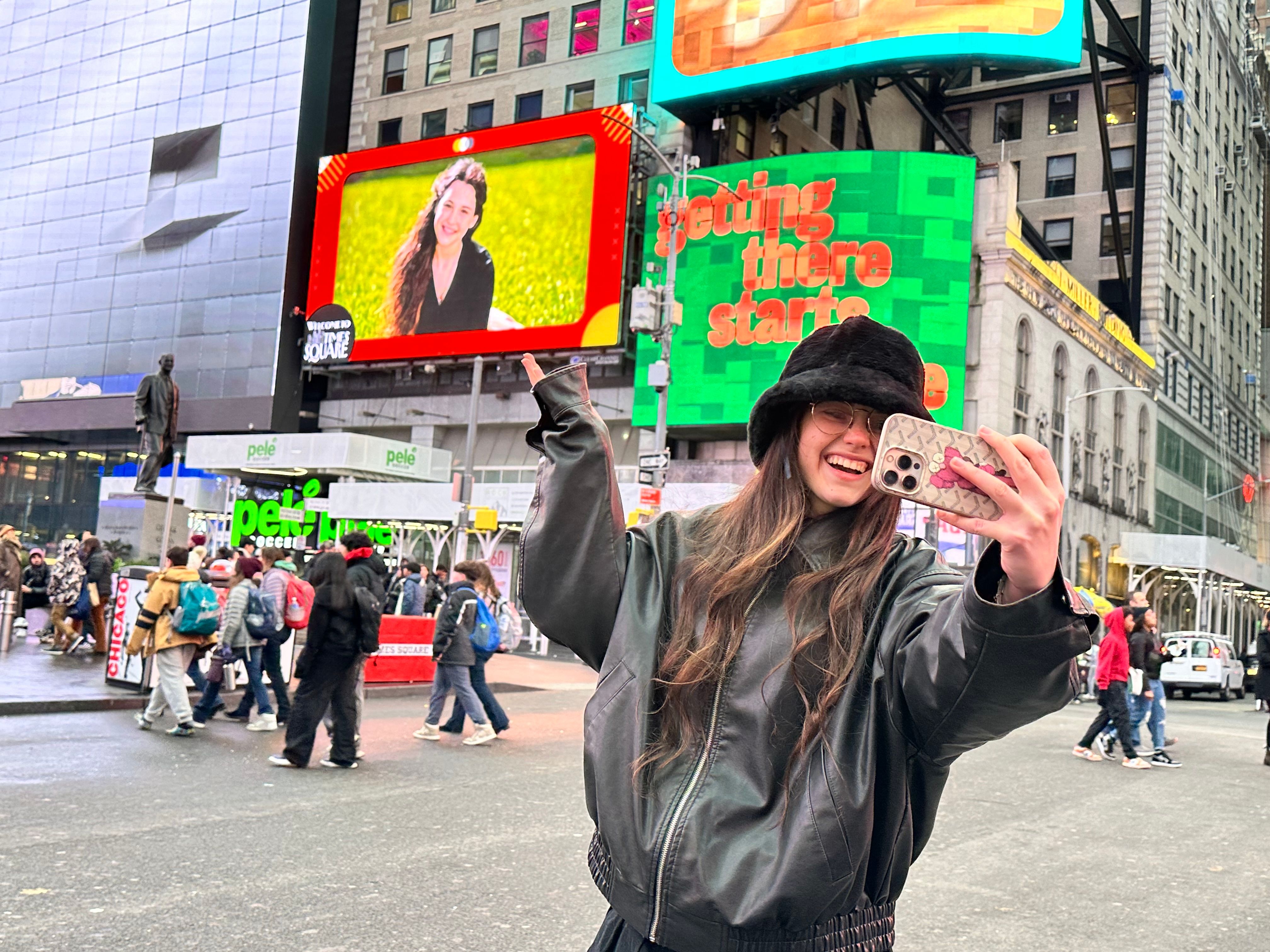 Smiling person taking a selfie in Times Square, NYC, wearing a black bucket hat and leather jacket with colorful digital billboards and a bustling crowd in the background.