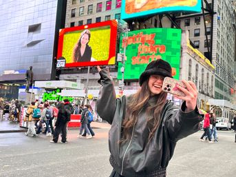 Smiling person taking a selfie in Times Square, NYC, wearing a black bucket hat and leather jacket with colorful digital billboards and a bustling crowd in the background.