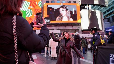 Smiling woman in a dark leather coat poses for a smartphone photo in crowded Times Square, NYC, beneath a giant LED billboard showing a portrait-style image.