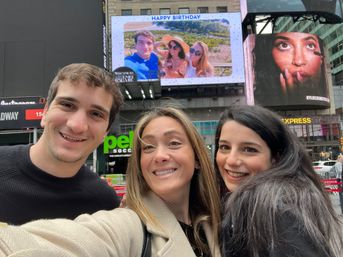 Smiling trio taking a selfie in Times Square, NYC, with a giant digital 'Happy Birthday' billboard and bright city ads behind them.