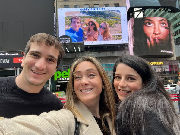 Smiling trio taking a selfie in Times Square, NYC, with a giant digital 'Happy Birthday' billboard and bright city ads behind them.