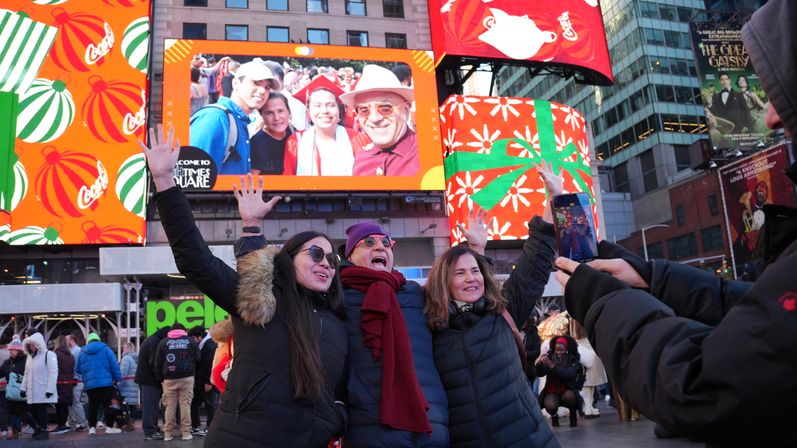 Three friends in winter coats cheer for a photo in Times Square, New York, beneath large colorful holiday billboards and a bustling city crowd.