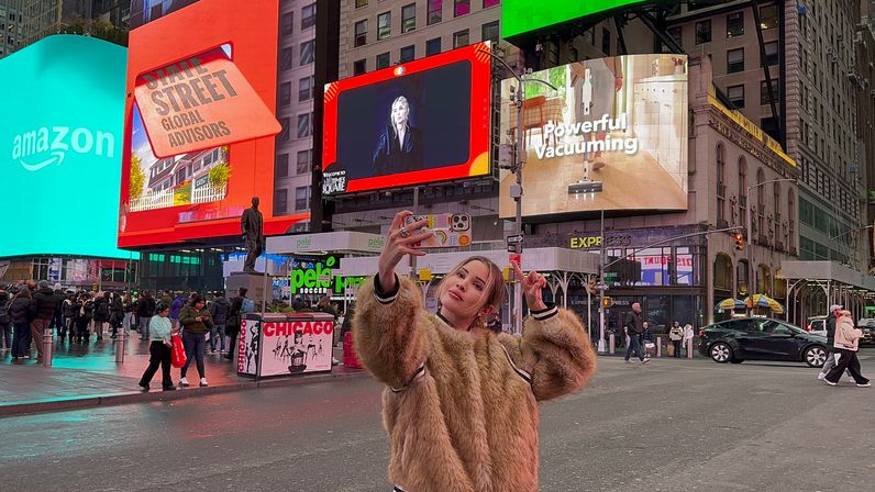 Person in a tan faux-fur coat taking a selfie and flashing a peace sign in Times Square, New York City, with bright LED billboards, crowds and traffic at dusk.
