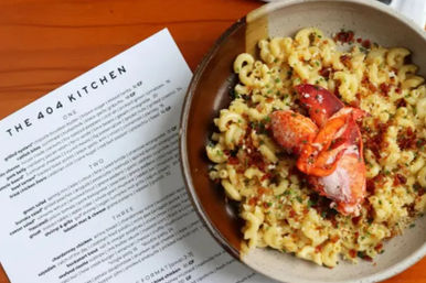 Overhead shot of a bowl of lobster mac and cheese with a lobster claw, crispy breadcrumb and herb garnish, pictured beside a restaurant menu on a wooden table