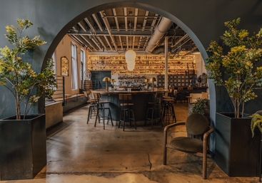 Industrial-chic bar interior framed by a large arched entry, featuring a central round wooden bar with stools, backlit shelves of bottles, exposed ceiling pipes, concrete floor, and potted plants for an inviting, urban lounge vibe.