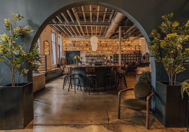 Industrial-chic bar interior framed by a large arched entry, featuring a central round wooden bar with stools, backlit shelves of bottles, exposed ceiling pipes, concrete floor, and potted plants for an inviting, urban lounge vibe.