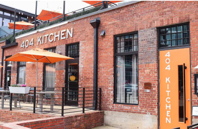 Downtown brick restaurant storefront with bright orange patio umbrellas, matching orange door, outdoor seating with metal railing and large industrial-style windows.