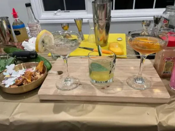 Three craft cocktails on a wooden tasting board at an at-home kitchen bar — two coupe glasses and a rocks glass garnished with lemon and cucumber, surrounded by citrus, cocktail tools and a snack bowl.