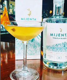 Sunny yellow tequila cocktail in a coupe glass with a citrus garnish, sitting beside a clear blanco tequila bottle with illustrated label and matching box on a polished bar counter