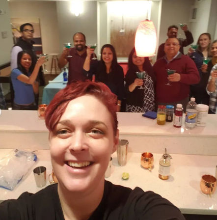 Smiling person taking a selfie at a lively home cocktail party, friends behind a kitchen bar raising teal martini glasses, with copper mugs and drink mixers on the counter.