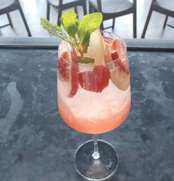 Refreshing pink plum spritz in a stemmed glass with crushed ice, plum slices and mint sprig, set on a dark bar counter with blurred chairs in the background