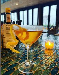 Amber cocktail in a coupe glass with a long lemon twist on a blue-green mosaic bar top, candlelit and large windows in a cozy modern bar setting
