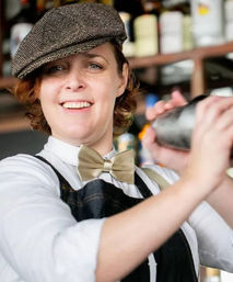 Smiling bartender in a flat cap and bow tie shaking a cocktail shaker behind a stocked bar in a cozy downtown cocktail spot.