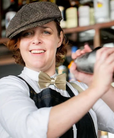 Smiling bartender in a flat cap and bow tie shaking a cocktail shaker behind a stocked bar in a cozy downtown cocktail spot.