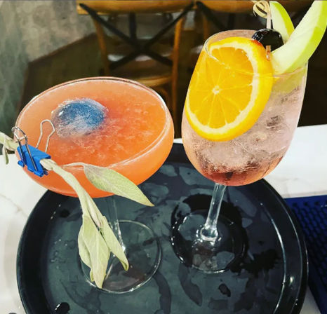 Two vibrant cocktails on a black serving tray at a bar — a frozen pink coupe with a blue ice sphere and clipped sage garnish, and a sparkling glass garnished with an orange slice, cherry, and apple wedge.