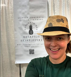 Smiling person in a straw fedora with pineapple print takes a selfie at an event booth featuring a pineapple logo banner promoting mixology, cocktail classes, and event services.