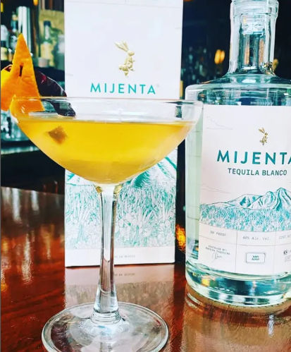 Zesty yellow tequila cocktail in a coupe glass with an orange peel garnish, pictured beside a clear tequila bottle and box on a polished bar countertop.