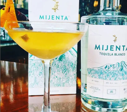 Zesty yellow tequila cocktail in a coupe glass with an orange peel garnish, pictured beside a clear tequila bottle and box on a polished bar countertop.