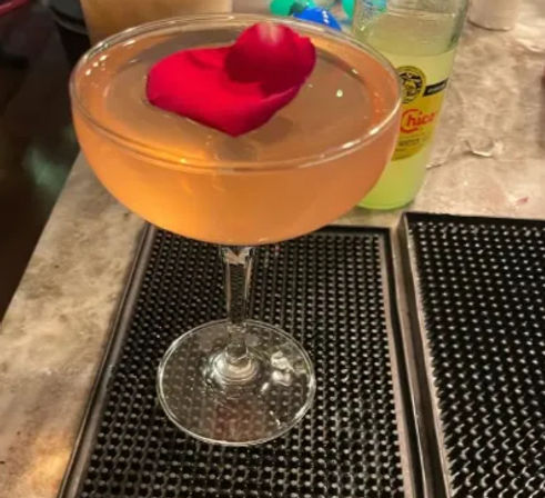 Peach-colored cocktail in a coupe glass topped with a single red rose petal, sitting on a black bar mat on a marble counter with a yellow soda bottle in the background.