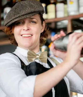 Smiling bartender mixing a cocktail with a shaker, wearing a flat cap, bow tie and apron in a lively cocktail bar with bottles on shelves