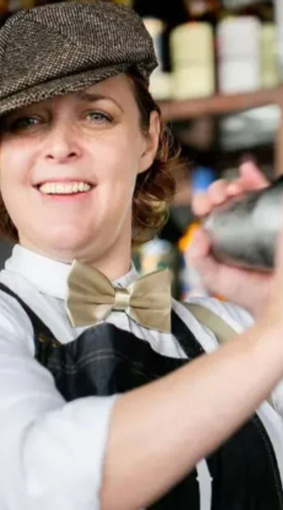 Smiling bartender mixing a cocktail with a shaker, wearing a flat cap, bow tie and apron in a lively cocktail bar with bottles on shelves