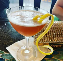 Frothy amber cocktail in a coupe glass with a long spiral lemon twist curling off the rim, served on a napkin on a bar top next to a sparkling clutch purse.