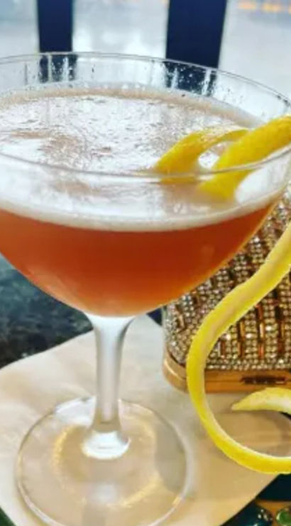 Frothy amber cocktail in a coupe glass with a long spiral lemon twist curling off the rim, served on a napkin on a bar top next to a sparkling clutch purse.