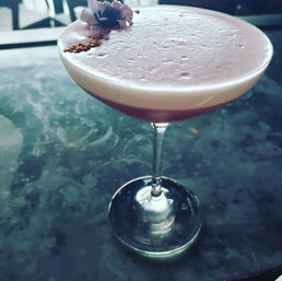 Pink frothy cocktail in a coupe glass topped with an edible flower and spice garnish, photographed on a marble bar top