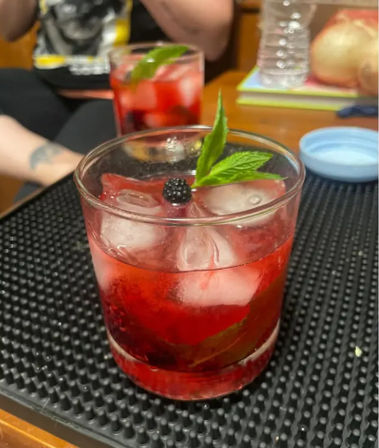 Vibrant red blackberry-mint cocktail with ice in a rocks glass on a black bar mat, garnished with a blackberry and mint sprig, second drink and blurred figure in the background.