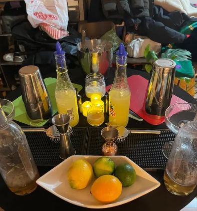 Home cocktail prep station with fresh lemons and limes in a white bowl, two yellow mixer bottles, metal cocktail shakers, jiggers, ice bucket and bar tools on a table — ready for citrus cocktails.