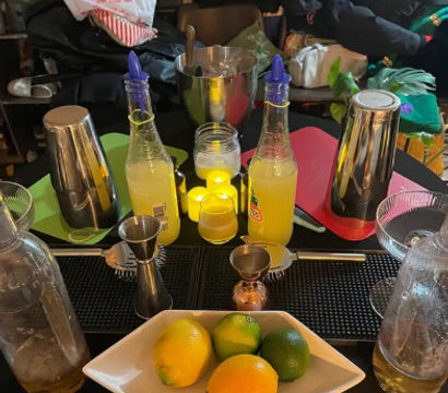Home cocktail prep station with fresh lemons and limes in a white bowl, two yellow mixer bottles, metal cocktail shakers, jiggers, ice bucket and bar tools on a table — ready for citrus cocktails.