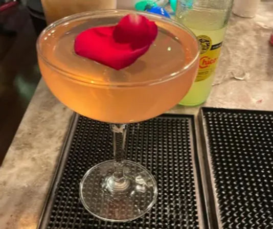 Peach-colored craft cocktail in a coupe glass topped with a single red rose petal resting on a bar counter mat