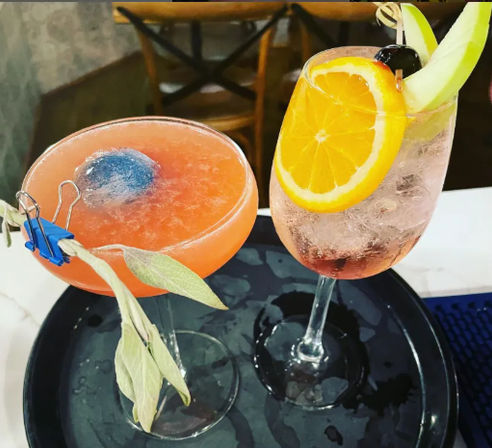 Two colorful cocktails on a black serving tray at a cocktail bar: a frosty pink-orange coupe with a blue ice sphere and sage sprig, and a sparkling wine glass garnished with an orange wheel, cherry, and melon slice.