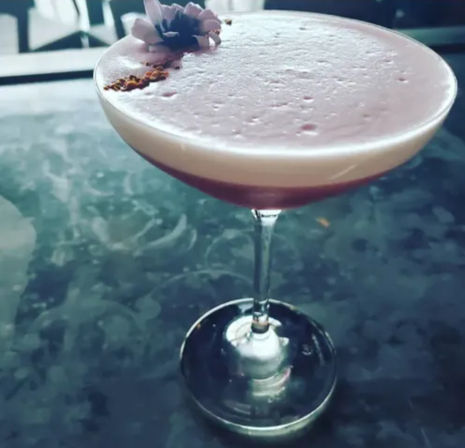 Frothy pink cocktail in a coupe glass, garnished with an edible flower and spice, resting on a dark marble bar surface.